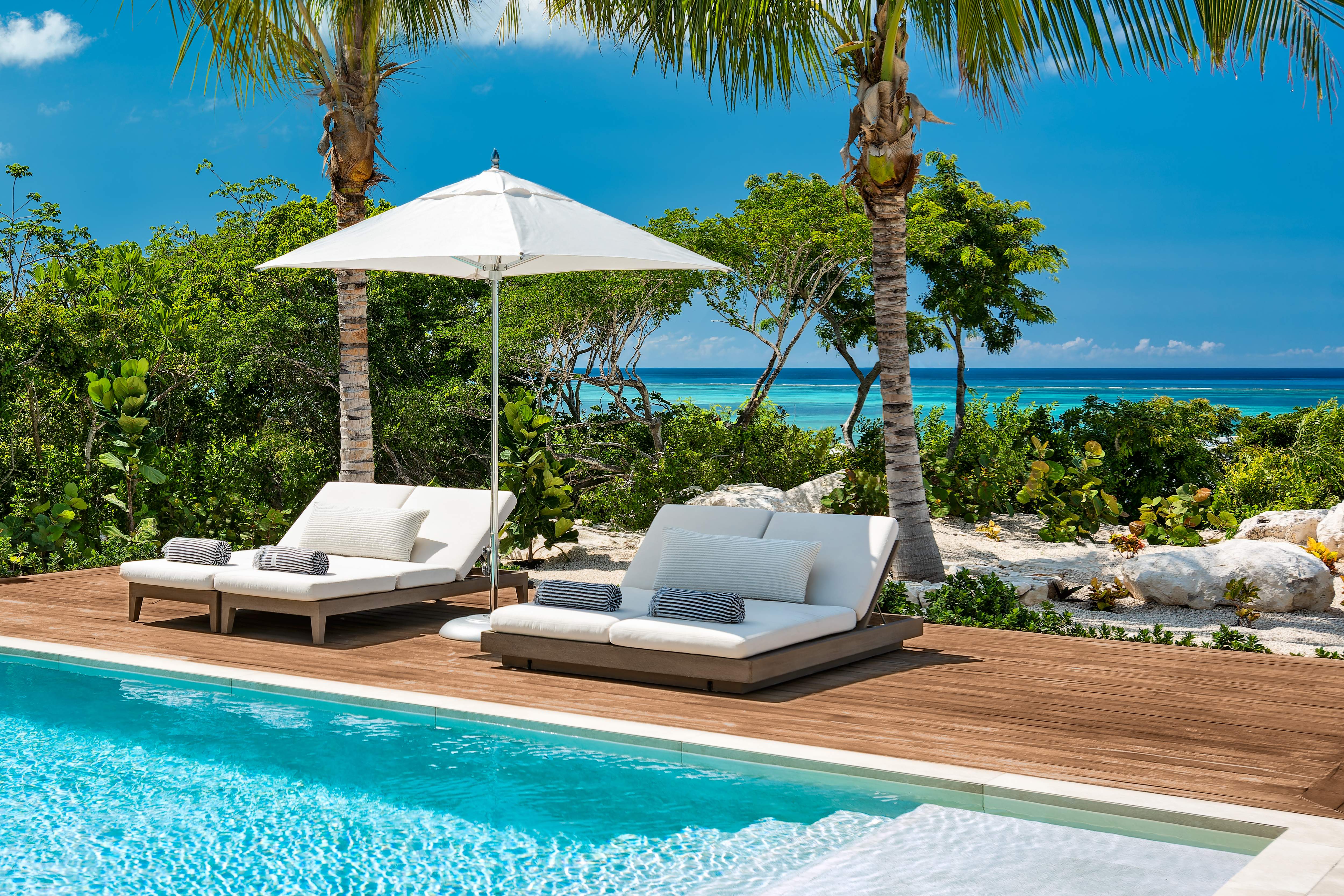 Poolside daybeds under a white umbrella with palms and turquoise ocean beyond at Beachwood Villa