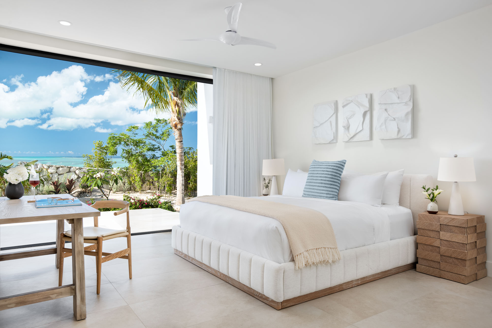 The Reef Suite at Beachwood Villa with king bed, oak nightstands, white textured wall art triptych, and a sliding glass wall opening to tropical gardens and partial ocean view