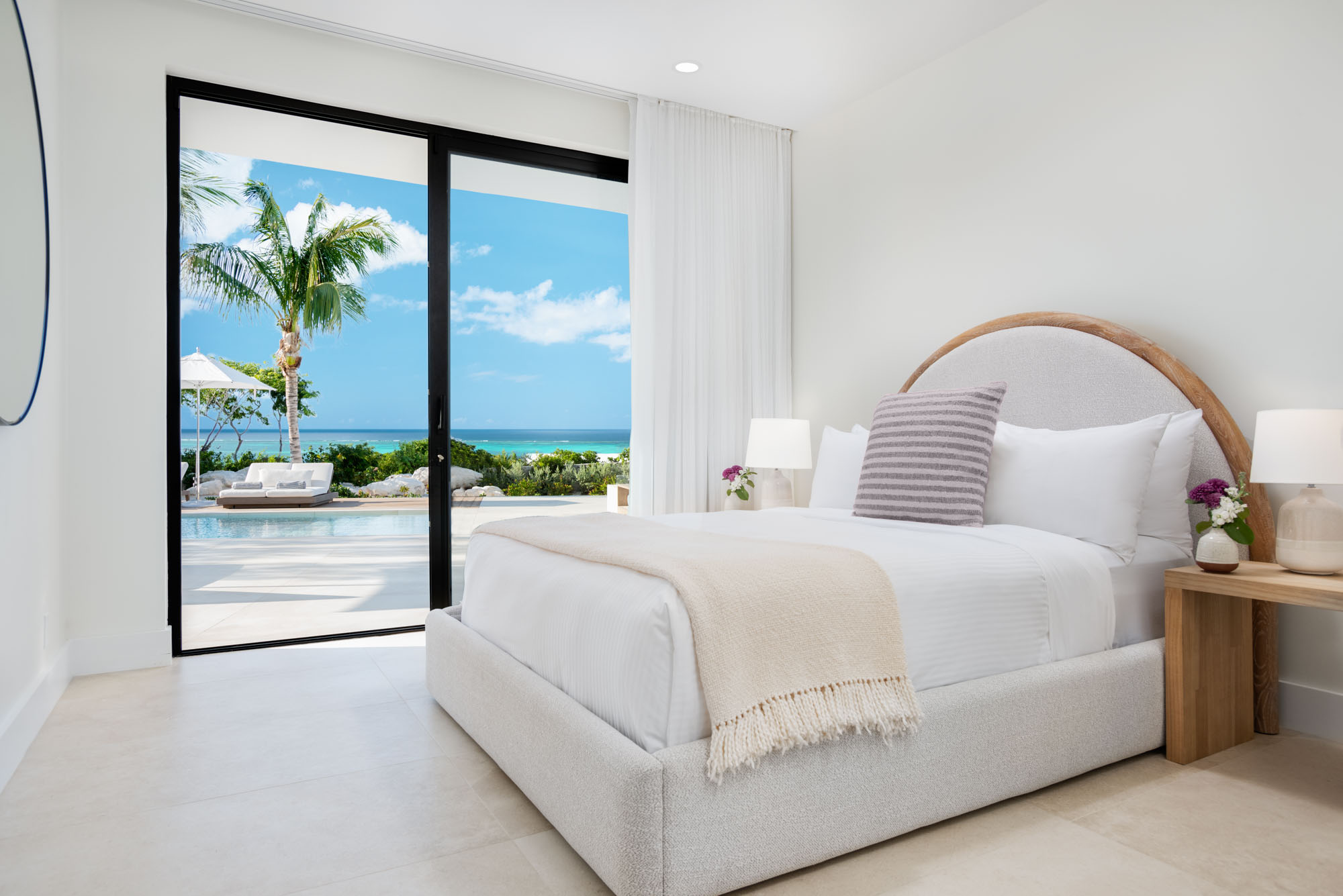 The Pool Suite at Beachwood Villa with queen bed, arched boucle headboard, white linens, and sliding glass wall opening directly to the pool deck and ocean view