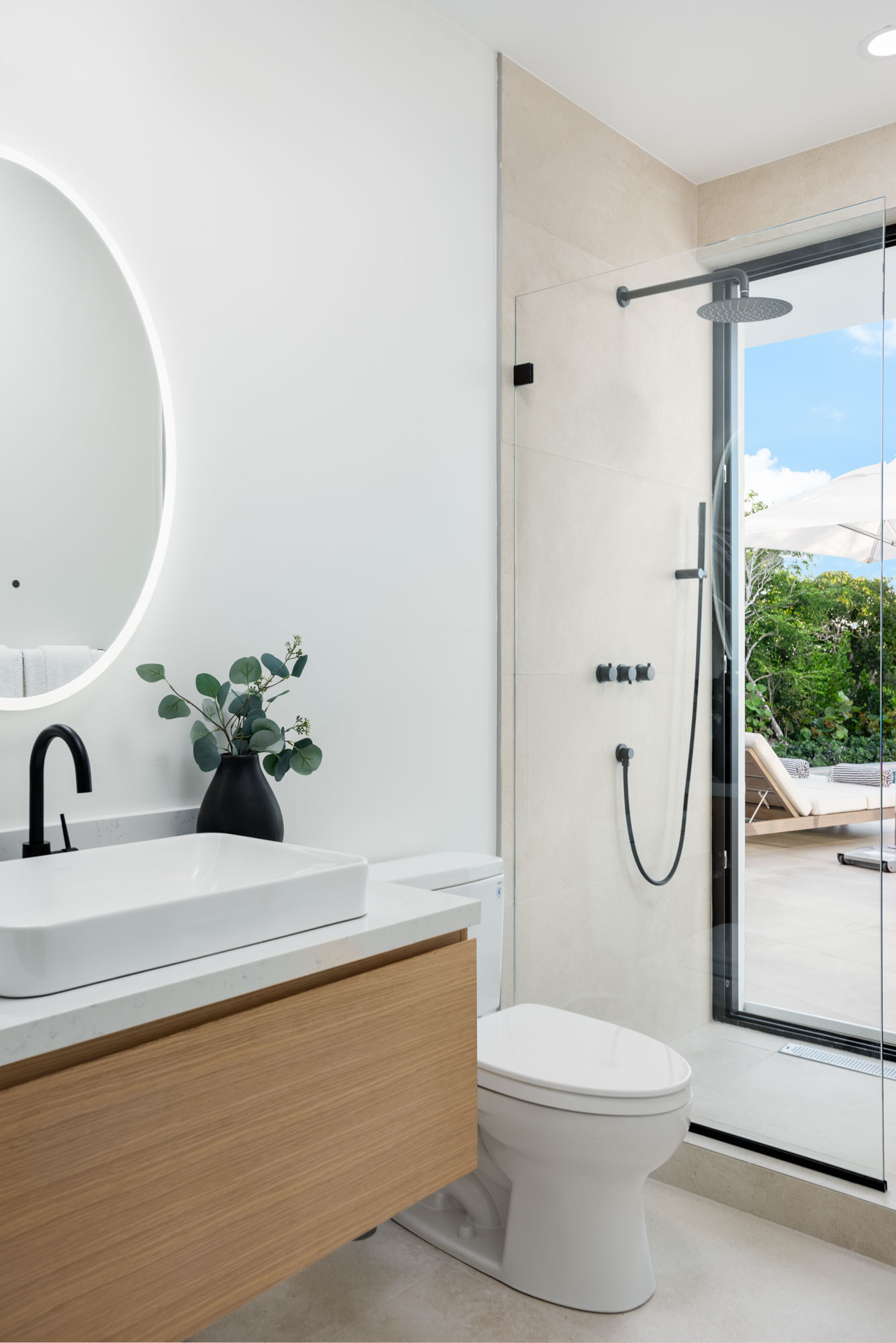 Garden Suite ensuite with floating oak vanity, vessel sink, round backlit mirror, and large glass-walled rain shower opening directly onto the pool deck