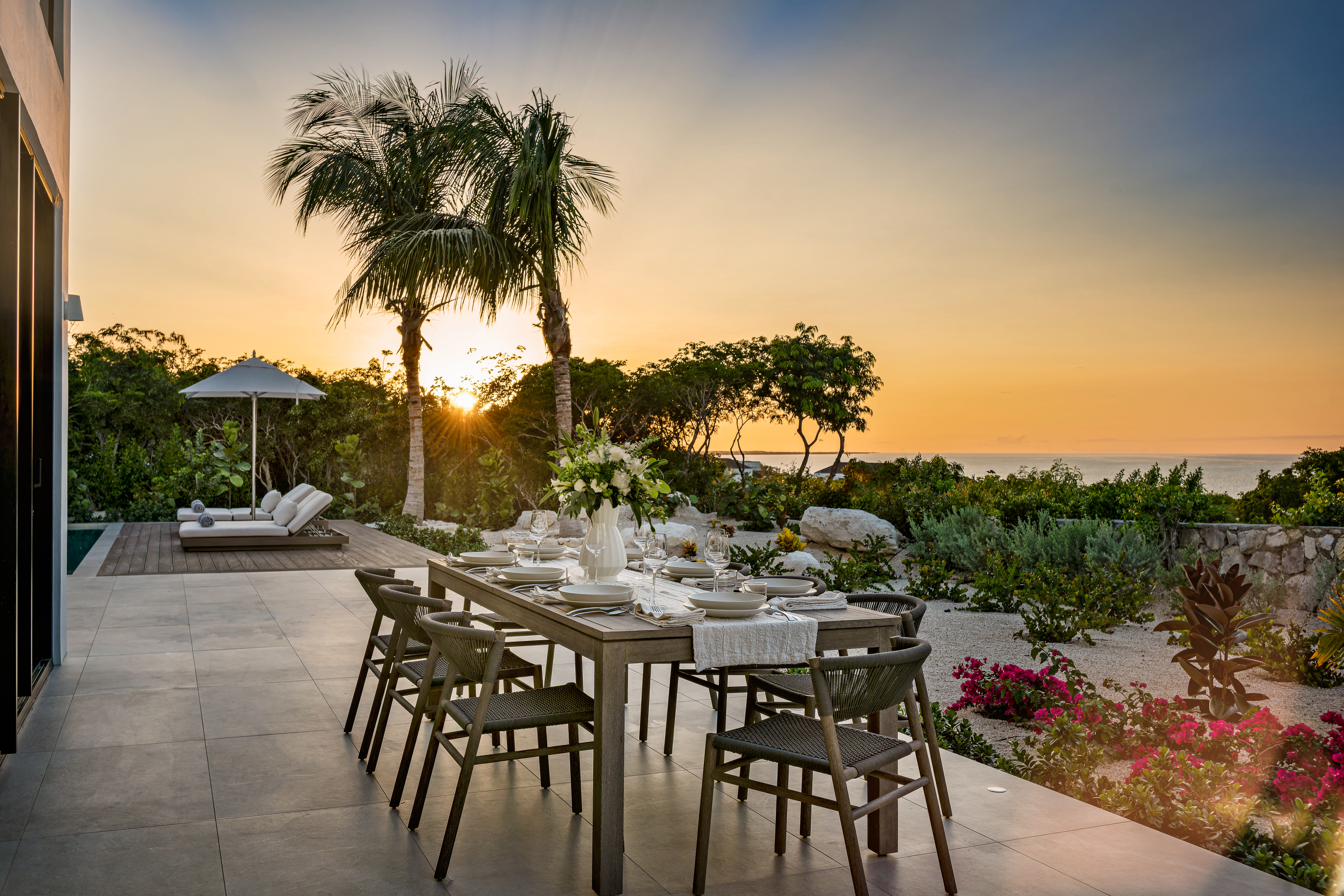 Long oak dining table set for sunset dinner at Beachwood Villa with palm trees silhouetted against the orange sky and the ocean horizon visible in Providenciales Turks and Caicos