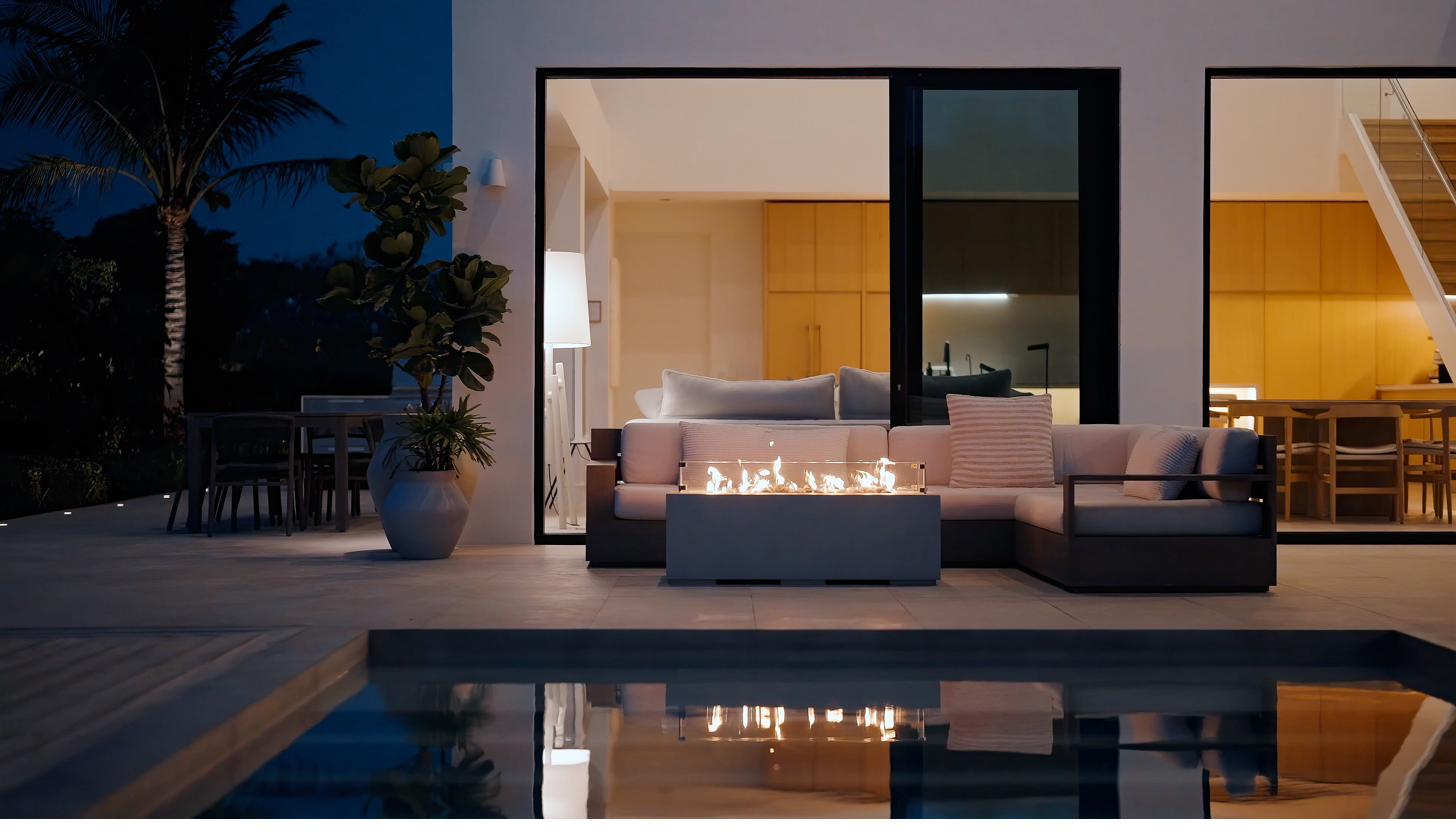 Beachwood Villa fire pit lit at night with the modular outdoor sofa, palms silhouetted against the dark sky, the villa glowing through glass walls behind, and the pool reflecting the firelight in Providenciales Turks and Caicos