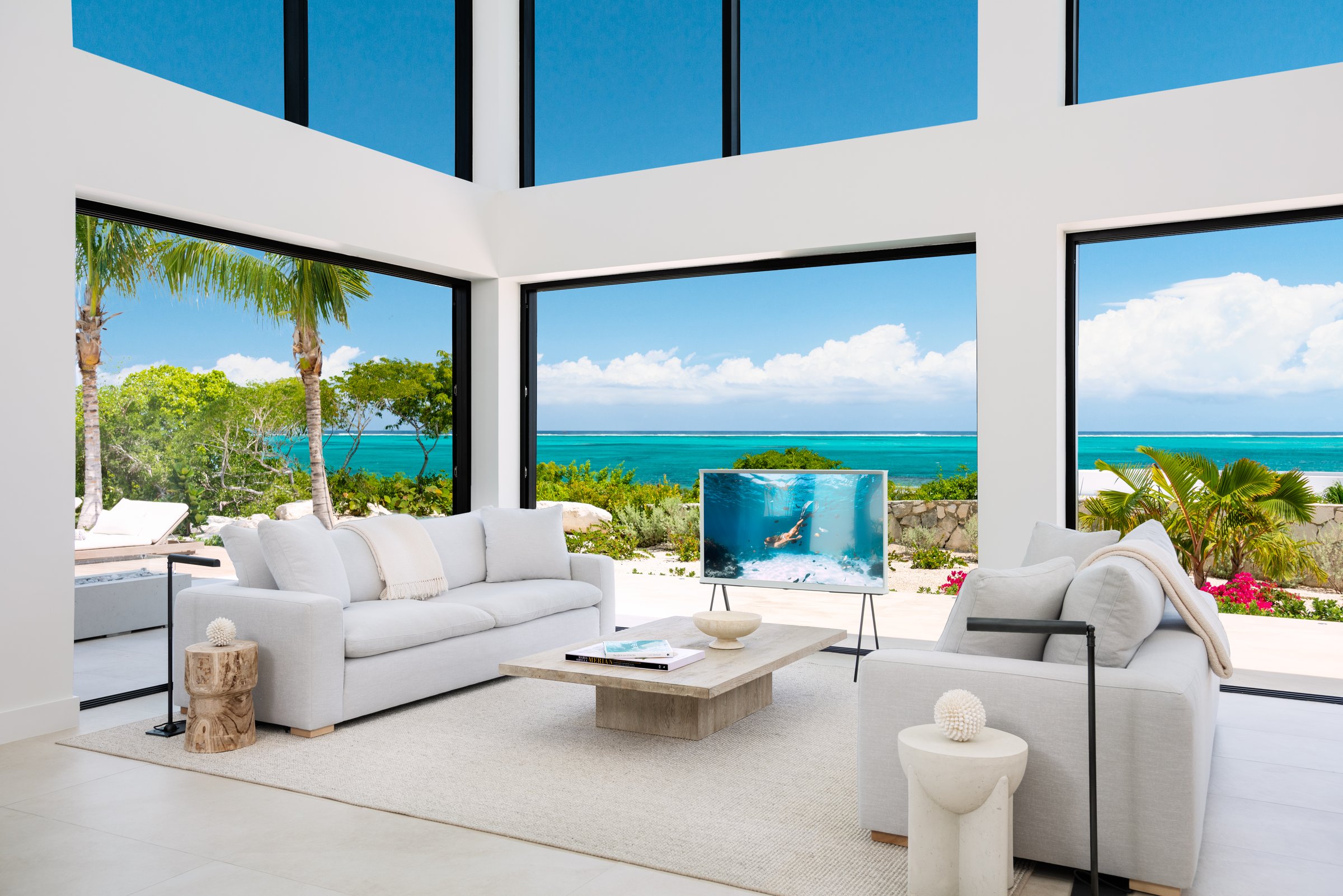 The great room at Beachwood Villa with round oak dining table styled for eight, two cream sofas, fourteen-foot kitchen island, twenty-foot ceilings, and floor-to-ceiling glass walls open to the turquoise Caribbean in Providenciales Turks and Caicos