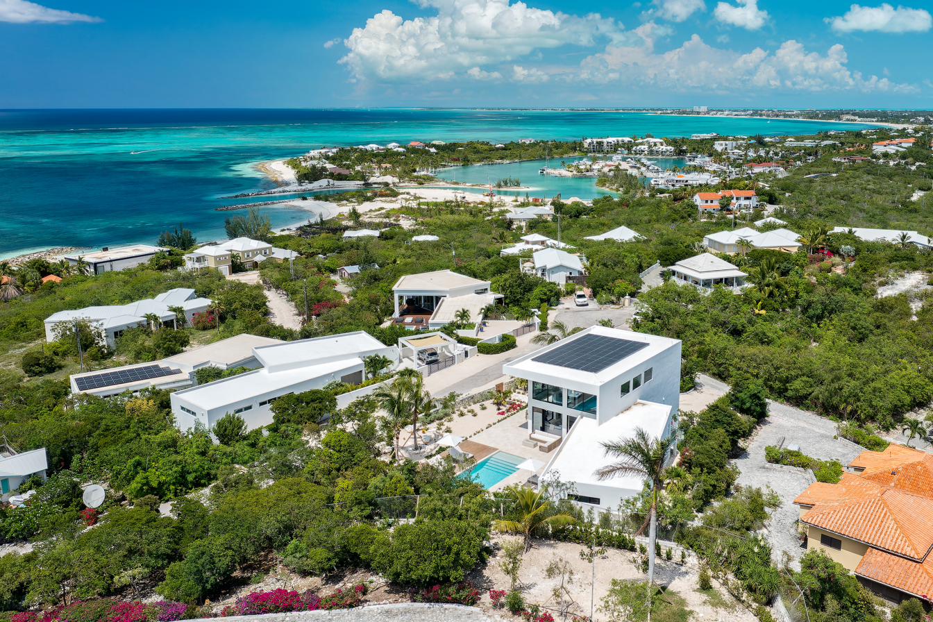 Aerial view from above Beachwood Villa showing the property in its elevated position with three bays of turquoise Caribbean water visible, Turtle Cove Marina in the middle distance, and the Providenciales coastline stretching out toward Grace Bay