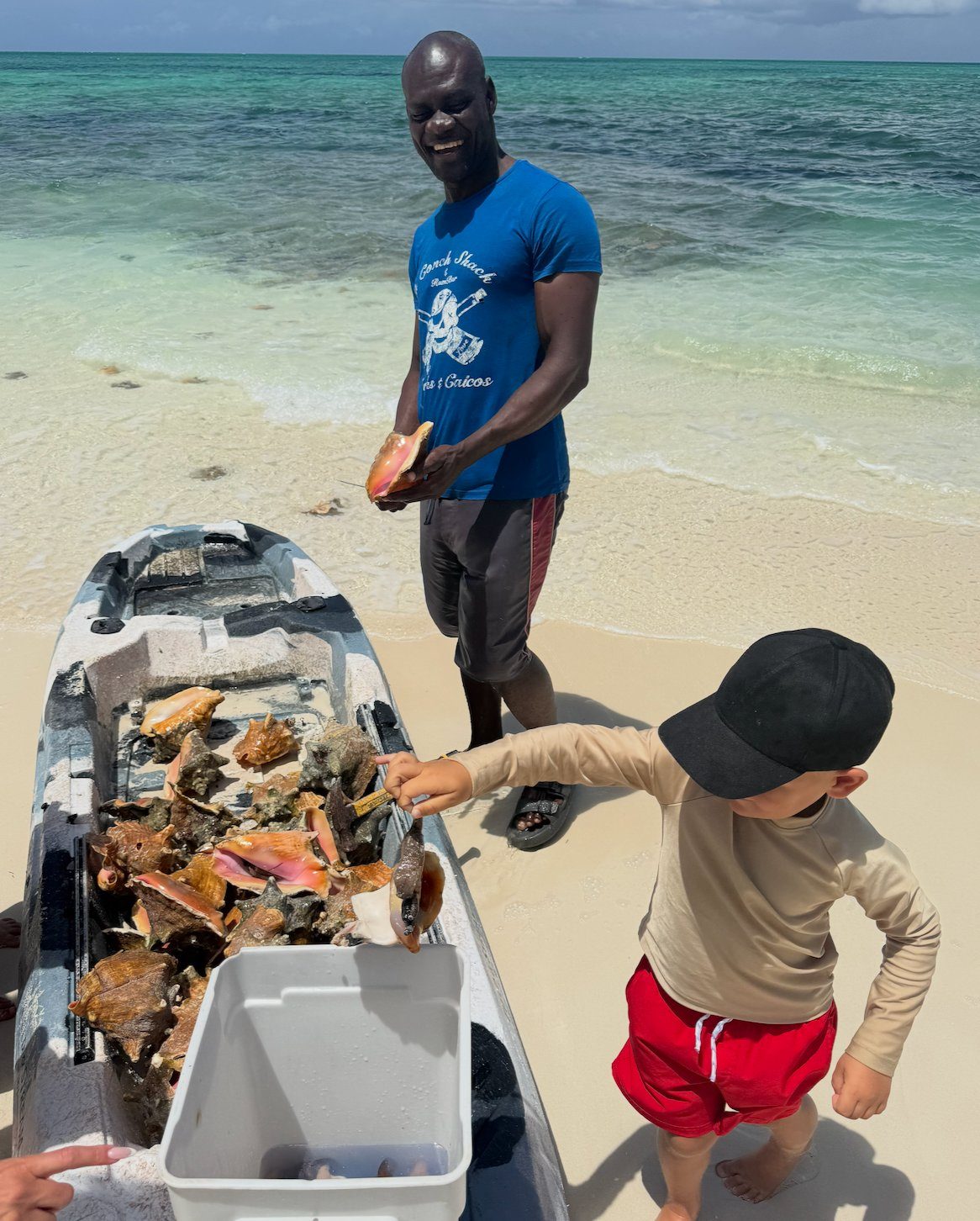 A man in a Conch Shack shirt holding fresh conch beside a kayak of conch shells on the beach, with a child watching, at da Conch Shack Providenciales