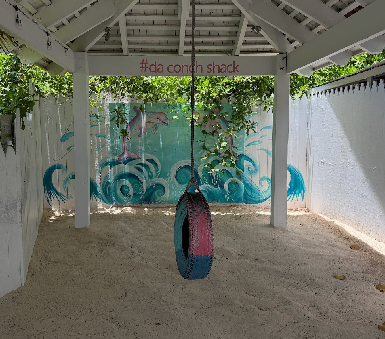 A pink and blue painted tire swing under a hashtag da conch shack sign with a dolphin mural, Providenciales